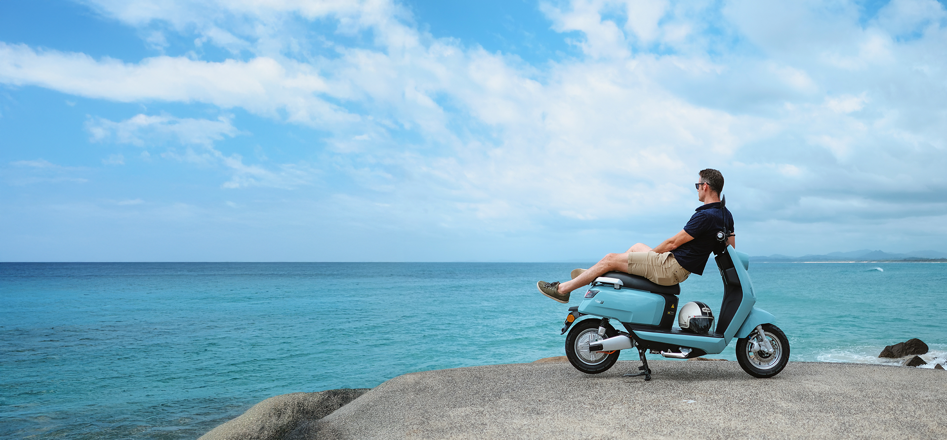 Man relaxes on a turquoise scooter at the edge of a rocky shore, overlooking blue ocean waves.